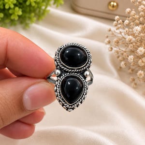May include: A silver ring featuring two smooth, black stones set in a vertical arrangement. The top stone is oval-shaped, while the bottom stone is pear-shaped. Both stones are framed by a twisted rope-style metal border with small silver beads.