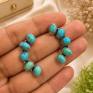 May include: A pair of ear climbers featuring five oval-shaped turquoise stones set in silver. Each stone is held by a scalloped silver bezel setting, arranged in a curved line to follow the shape of the earlobe. The stones display natural blue and green hues with dark matrix veining.