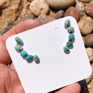 May include: A pair of ear climber earrings featuring multiple oval-shaped turquoise stones set in silver. Each earring consists of five individual stones arranged in a curved line, showcasing natural blue and green hues with dark matrix patterns.