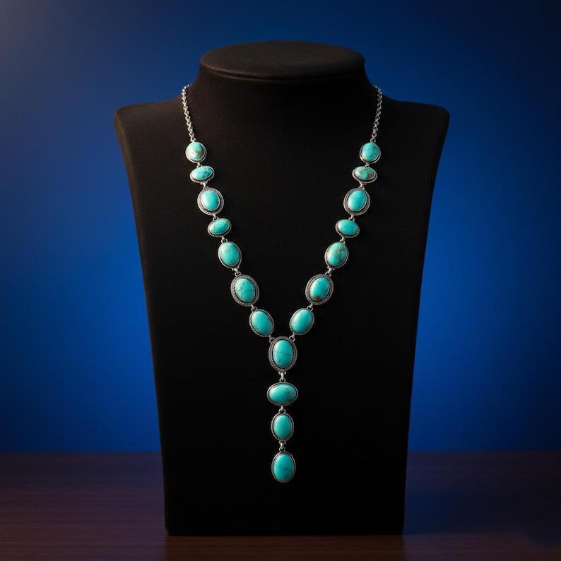 May include: A silver necklace featuring oval turquoise stones. The necklace is displayed on a black mannequin. The stones are set in silver metal and linked by a fine chain. The necklace has a Y-shaped design.