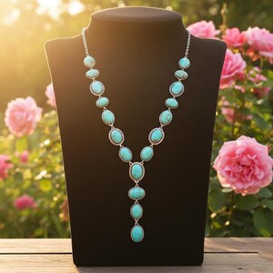 May include: A silver necklace with oval turquoise stones displayed on a black mannequin. The necklace has a Y-shaped pendant design, with the stones set in silver metal. The background features pink roses and sunlight.