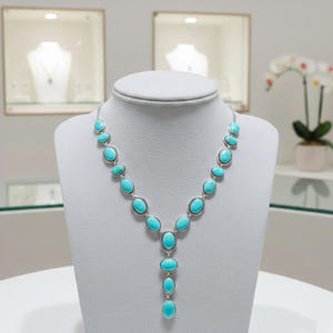 May include: A silver necklace featuring oval turquoise stones. The stones are set in a silver frame and arranged in a cascading design. The necklace is displayed on a white bust, highlighting its elegant design.