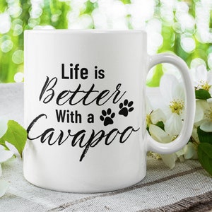 May include: White ceramic coffee mug with black text that reads "Life is Better With a Cavapoo" and two paw prints. The mug is sitting on a white and brown surface with white flowers.