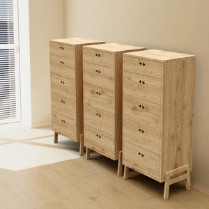 Puede incluir: Tres cómodas de madera clara, cada una con seis cajones y un acabado de veta clara. Tienen pequeños tiradores oscuros y están sobre patas cortas. La habitación está iluminada por la luz natural de una ventana.