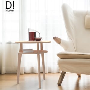 Könnte beinhalten: Ein heller Holz-Beistelltisch mit runder Tischplatte und einem unteren Regal. Der Tisch hat vier Beine und steht neben einem weißen Sessel.