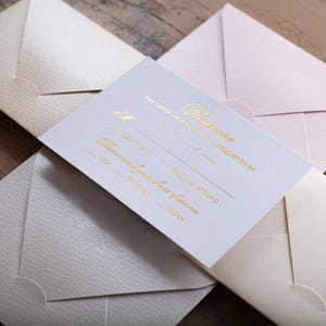 A white RSVP card with gold foil text is centered among four textured envelopes in cream, light pink, and light gray, on a wooden surface.