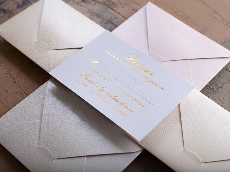 A white RSVP card with gold foil text is centered among four textured envelopes in cream, light pink, and light gray, on a wooden surface.