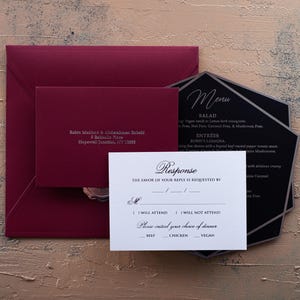 A white RSVP card with embossed text, a dark red envelope, and a black menu card are arranged on a textured brown and blue surface.