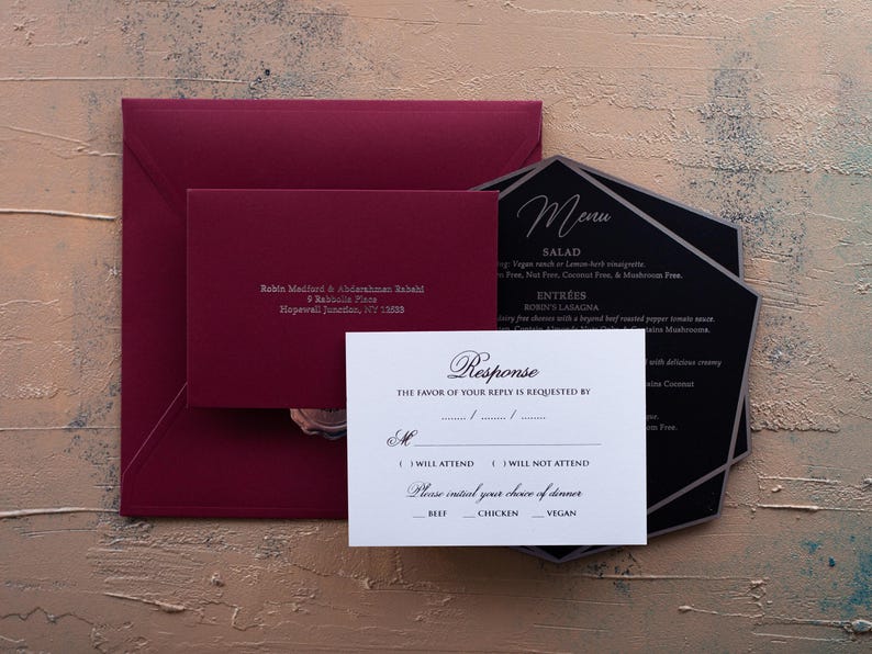 A white RSVP card with embossed text, a dark red envelope, and a black menu card are arranged on a textured brown and blue surface.