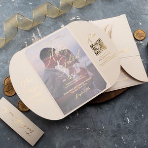 Puede incluir: Una invitación de boda plegada con una foto de la pareja en el frente. La invitación es de color crema con letras doradas y un código QR. El texto de la invitación dice "Ashley & Gabriel, Vie 25, Veinte Veinte-Tres, a las 6:00 PM, Sunflower Restaurant & Resort, 9999 River Bloom Lane, Recepción a seguir."