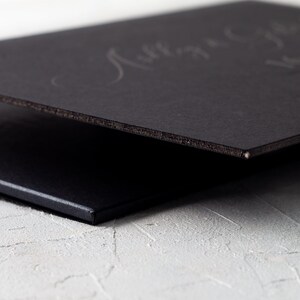 A close-up side view of a thick black wedding invitation card with a black core, showing the edge and a subtle embossed design on the surface.