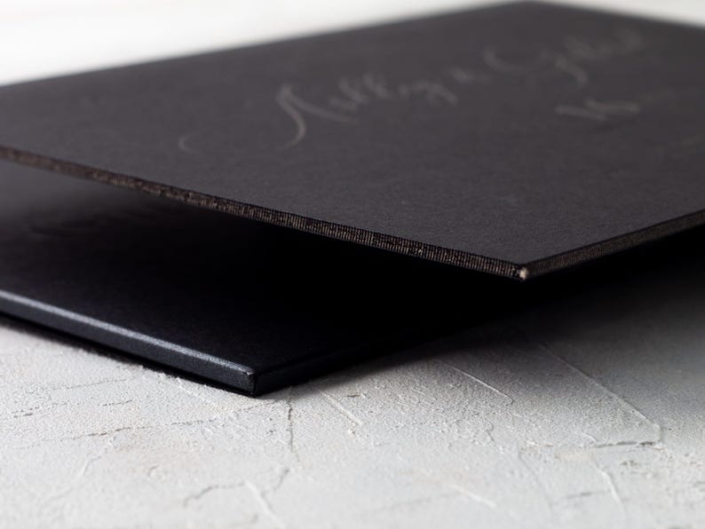 A close-up side view of a thick black wedding invitation card with a black core, showing the edge and a subtle embossed design on the surface.