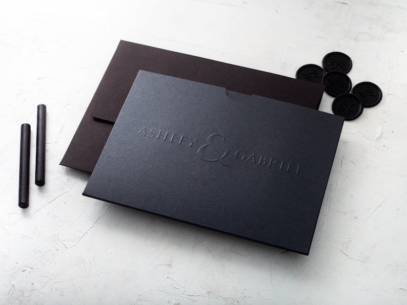 May include: A dark navy blue box embossed with the names "Ashley & Gabriel" is shown, alongside a dark brown envelope and several black wax seals. Two black sealing sticks are also present, suggesting a stationery or invitation theme.