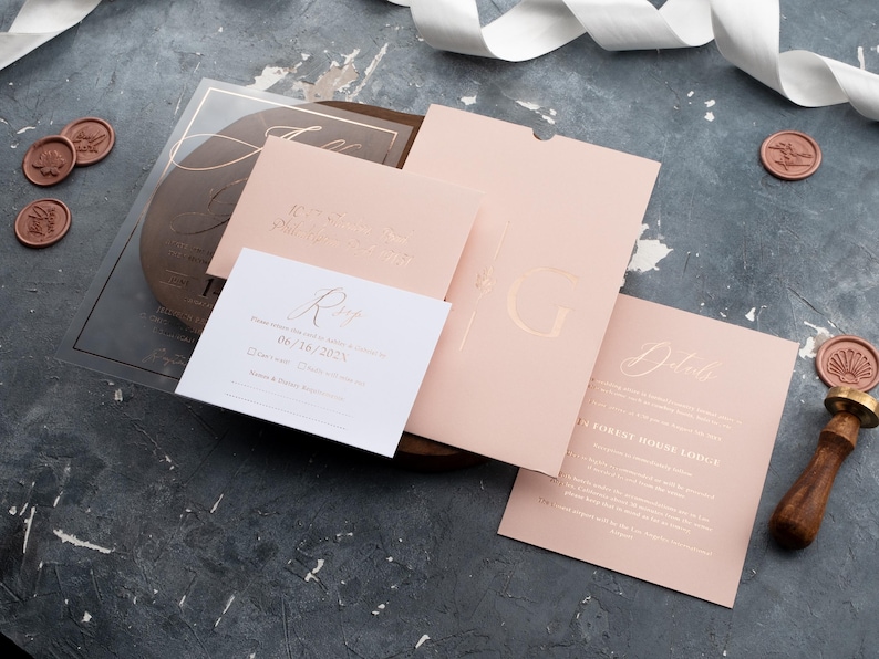 Blush pink wedding stationery flat lay with a clear acrylic invitation, sleeve envelope and optional printed RSVP and details cards.