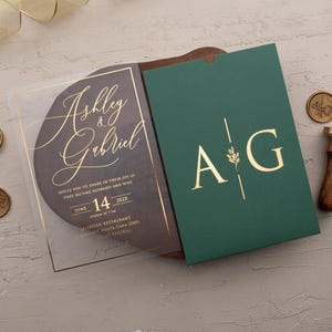 Personalized clear acrylic wedding invitation with rose gold foil beside an emerald green sleeve envelope with monogram.