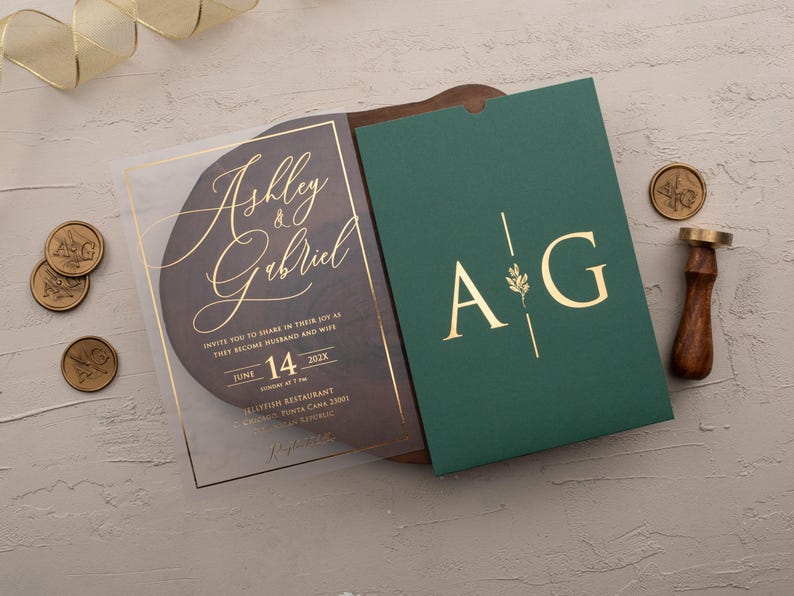 Personalized clear acrylic wedding invitation with rose gold foil beside an emerald green sleeve envelope with monogram.