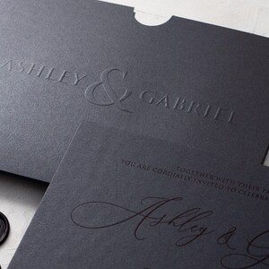 A black wedding invitation with copper script and a black envelope with embossed names are shown at an angle on a white textured surface.