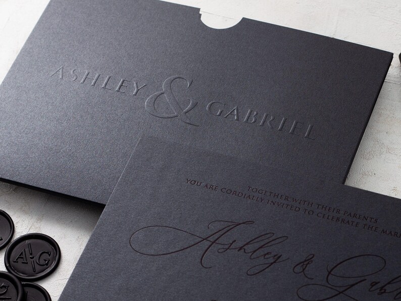 A black wedding invitation with copper script and a black envelope with embossed names are shown at an angle on a white textured surface.