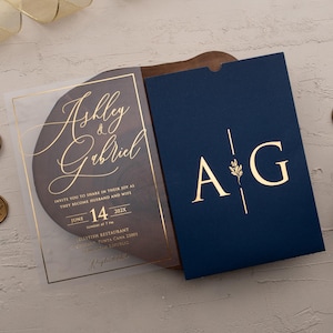 Personalized clear acrylic wedding invitation with rose gold foil beside a navy blue sleeve envelope with monogram.