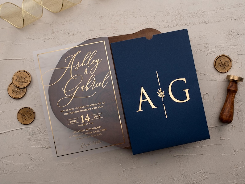 Personalized clear acrylic wedding invitation with rose gold foil beside a navy blue sleeve envelope with monogram.