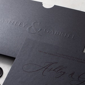 May include: Dark grey wedding invitation set with embossed names "Ashley & Gabriel". The invitation includes a matching envelope and a smaller card with elegant script. Black wax seals with initials are visible.