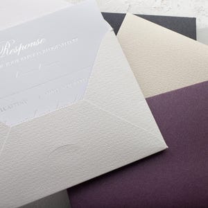A light gray RSVP card with embossed text is partially inserted into a light gray envelope, with a cream and a dark purple envelope visible behind it.