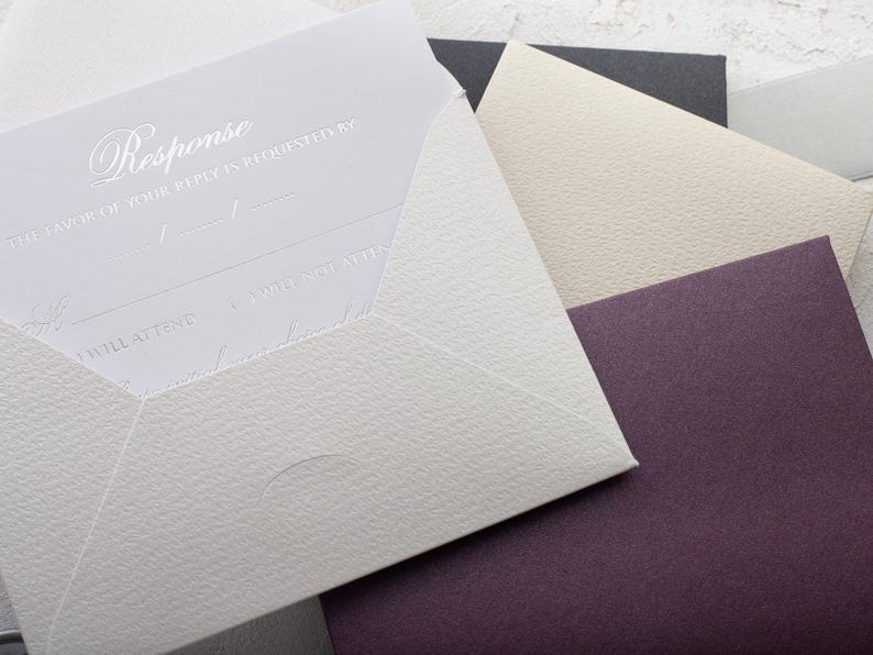 A light gray RSVP card with embossed text is partially inserted into a light gray envelope, with a cream and a dark purple envelope visible behind it.