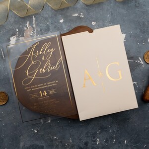 Personalized clear acrylic wedding invitation with rose gold foil beside an ivory sleeve envelope with monogram.