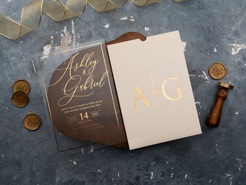 Personalized clear acrylic wedding invitation with rose gold foil beside an ivory sleeve envelope with monogram.
