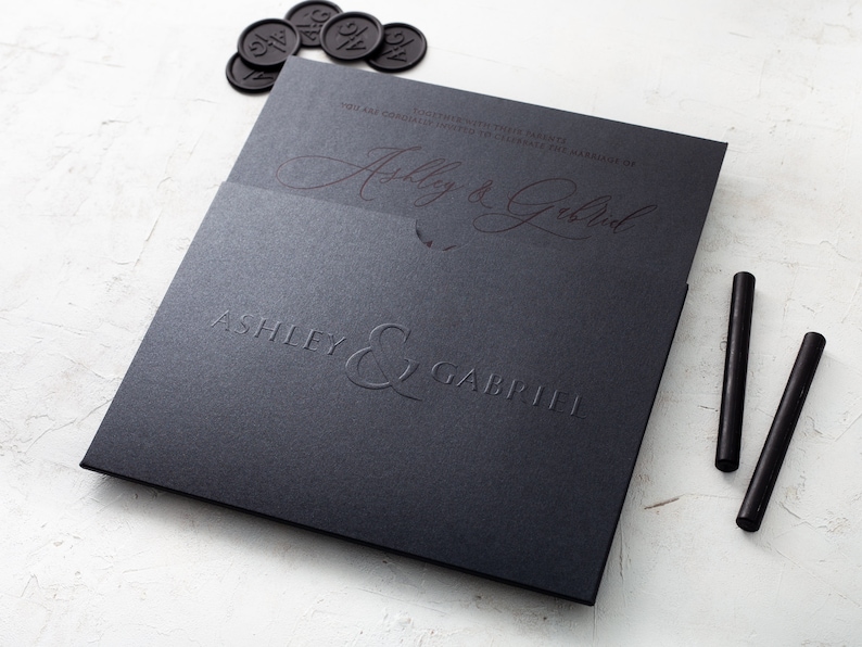 May include: A black wedding invitation featuring the names "Ashley & Gabriel" in embossed lettering. The invitation is presented with black wax seals and two black sealing sticks. The design is elegant and minimalist.