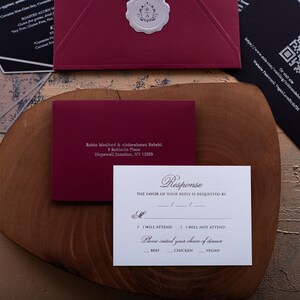 A white RSVP card with embossed text, a dark red envelope with a printed address, and a black card with a QR code are arranged on a wooden board.