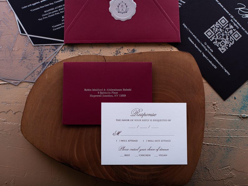 A white RSVP card with embossed text, a dark red envelope with a printed address, and a black card with a QR code are arranged on a wooden board.