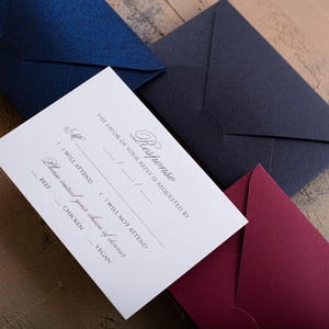A white RSVP card with embossed text is centered among three envelopes in dark blue, navy, and dark red, all on a textured brown and blue surface.