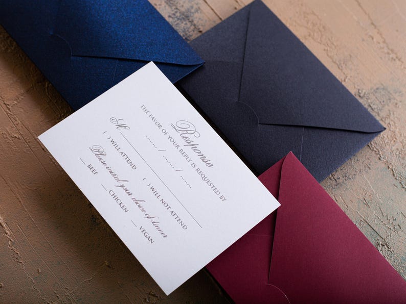 A white RSVP card with embossed text is centered among three envelopes in dark blue, navy, and dark red, all on a textured brown and blue surface.