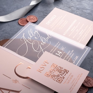 Angled view of a clear acrylic wedding invitation, blush pink sleeve envelope and optional rose gold foil QR RSVP and details cards.
