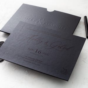 A black wedding invitation with "Ashley & Gabriel" embossed in copper script, and a matching black envelope with "Ashley & Gabriel" embossed in black, rests on a white textured surface next to black wax seals and wax sticks.