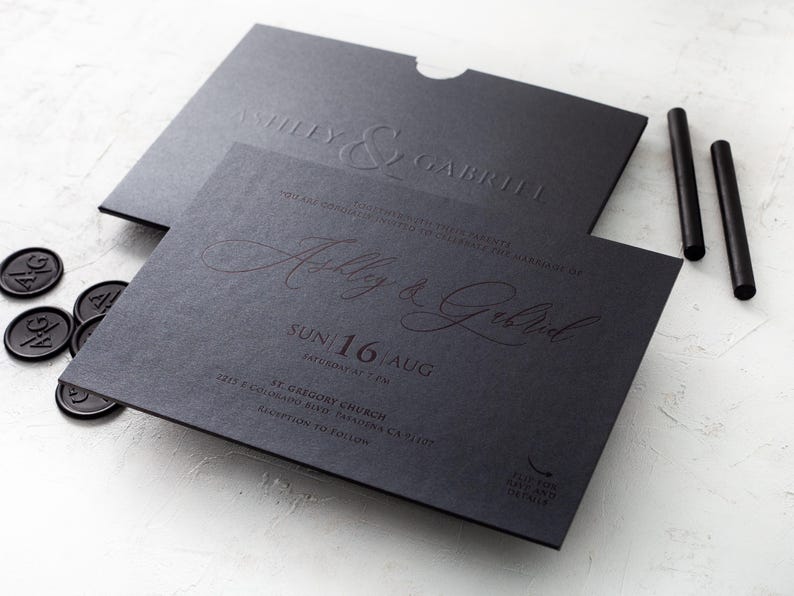 A black wedding invitation with "Ashley & Gabriel" embossed in copper script, and a matching black envelope with "Ashley & Gabriel" embossed in black, rests on a white textured surface next to black wax seals and wax sticks.