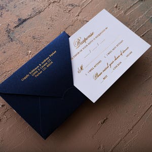 A cream-colored RSVP card with gold foil text is partially inserted into a dark blue envelope with a printed return address, all on a textured brown and blue surface.