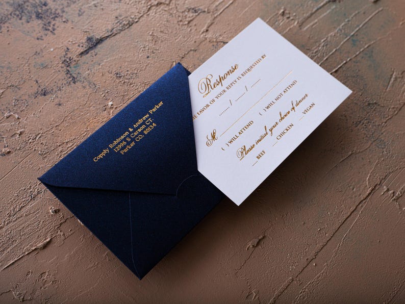 A cream-colored RSVP card with gold foil text is partially inserted into a dark blue envelope with a printed return address, all on a textured brown and blue surface.