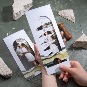 May include: White wedding invitation with a photo of a couple, featuring torn paper design elements. The invitation includes text and is held by a person. Decorative wax seals and stone fragments are also present.