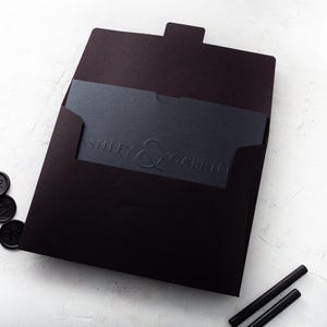 A black wedding invitation with embossed names is partially inserted into a black envelope, surrounded by black wax seals and wax sticks on a white textured surface.