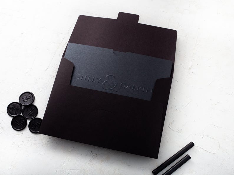 A black wedding invitation with embossed names is partially inserted into a black envelope, surrounded by black wax seals and wax sticks on a white textured surface.