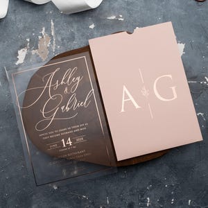 Clear acrylic wedding invitation with rose gold foil lettering beside a blush pink monogram sleeve envelope on a wooden tray.