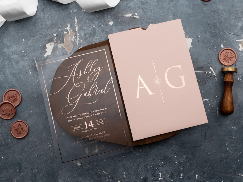 Clear acrylic wedding invitation with rose gold foil lettering beside a blush pink monogram sleeve envelope on a wooden tray.