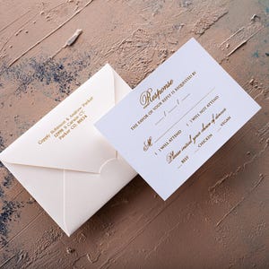 A cream-colored RSVP card with gold foil text and a matching envelope with a printed return address are displayed on a textured brown and blue surface.