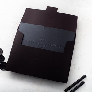May include: A black wedding invitation suite. It includes a dark envelope, a card with embossed text reading "ASHLEY & GABRIEL", and black wax seals. Two black sealing sticks are also present.