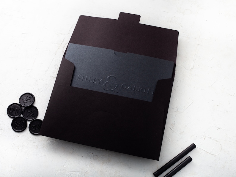 May include: A black wedding invitation suite. It includes a dark envelope, a card with embossed text reading "ASHLEY & GABRIEL", and black wax seals. Two black sealing sticks are also present.