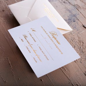 A cream-colored RSVP card with gold foil text is in the foreground, with a matching envelope with a printed return address blurred in the background, all on a textured brown and blue surface.