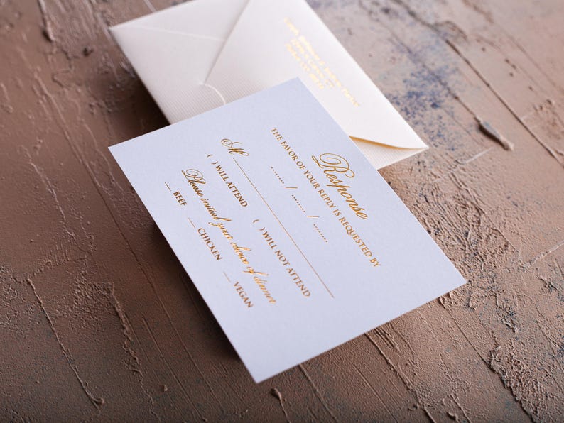 A cream-colored RSVP card with gold foil text is in the foreground, with a matching envelope with a printed return address blurred in the background, all on a textured brown and blue surface.