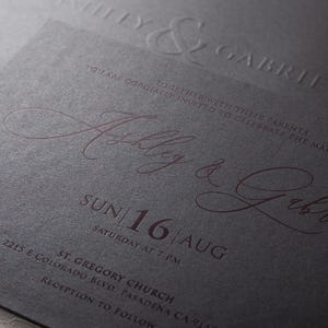 A close-up of a black wedding invitation with "Ashley & Gabriel" embossed in copper script, along with the date "SUN 16 AUG" and event details, on a textured surface.